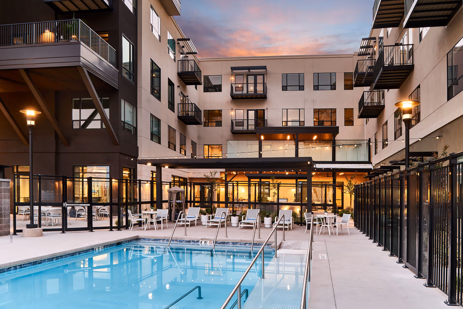 Outdoor swimming pool area at a senior living facility during sunset, surrounded by a black metal fence. There are several white lounge chairs and tables with chairs arranged around the pool. The building in the background has multiple balconies and large windows, with warm lights glowing from inside. String lights are hung above the seating area near the building.