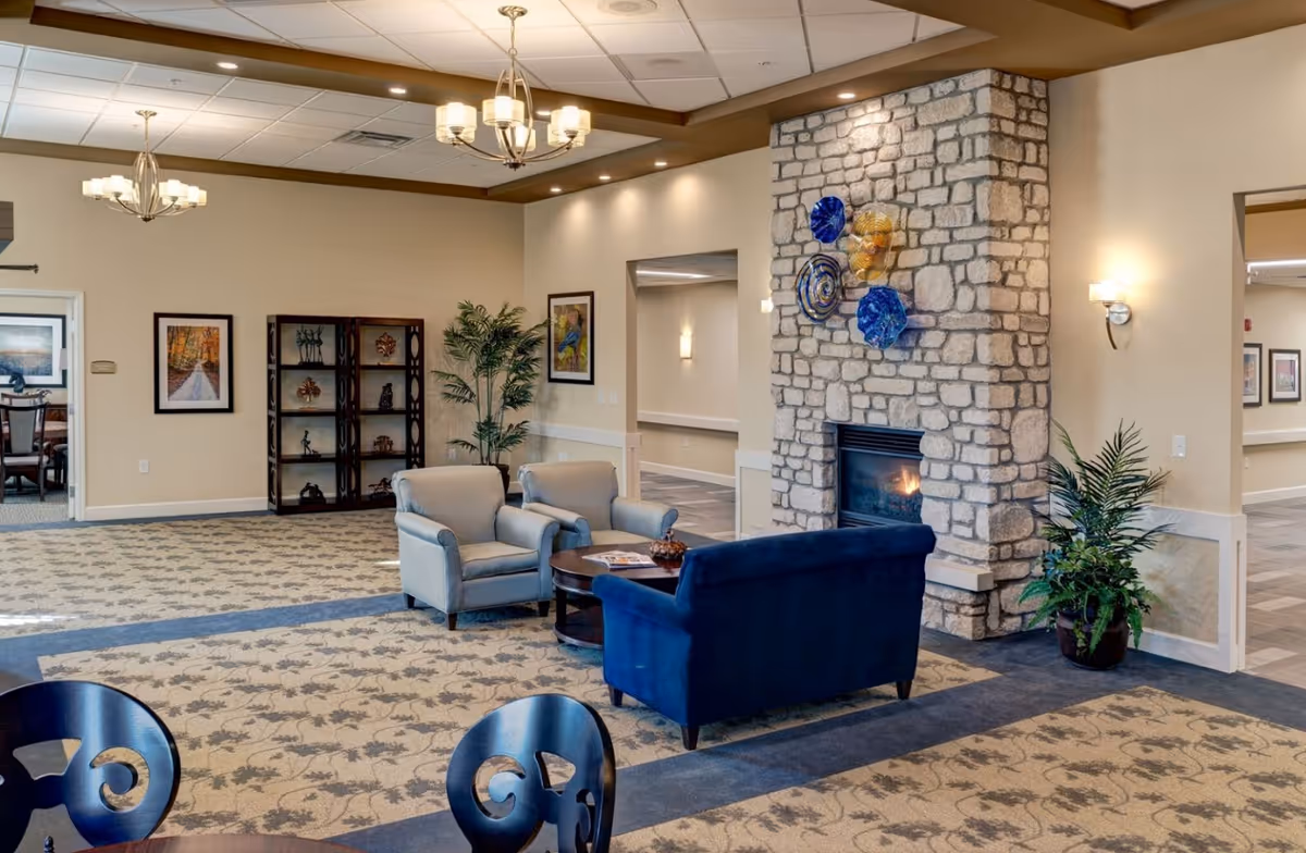 A cozy senior living facility common area with a stone fireplace decorated with colorful glass art. The room features a blue loveseat, two beige armchairs, a round wooden coffee table, and patterned carpet. There are plants, framed artwork on the walls, and two chandeliers hanging from the ceiling.