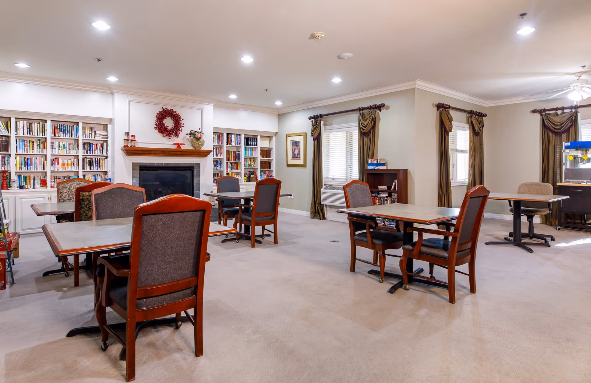 A spacious common room with several tables and chairs arranged for seating. The room features built-in bookshelves filled with books, a fireplace decorated with a red wreath, and windows with brown curtains. There is also a popcorn machine in the corner and framed artwork on the walls.