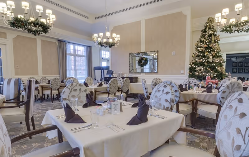 Elegantly decorated dining room with set tables and patterned chairs, chandeliers overhead and a decorated Christmas tree.