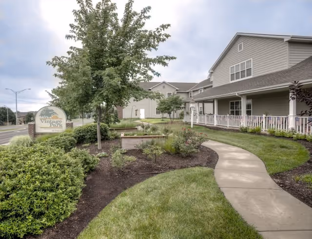 Exterior view of Vintage Park at Stanley senior living facility showing a landscaped garden with bushes, trees, and a curved concrete walkway leading to the building entrance. The building has gray siding, white trim, and a covered porch with white railings. A sign with the facility name is visible near the road.