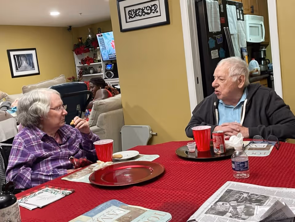 Two elderly individuals sitting at a table covered with a red tablecloth, engaged in conversation. The table has red plastic cups, a water bottle, a soda can, and some plates with food. In the background, there is a living room area with a woman sitting on a couch, a TV, and some decorations on shelves.