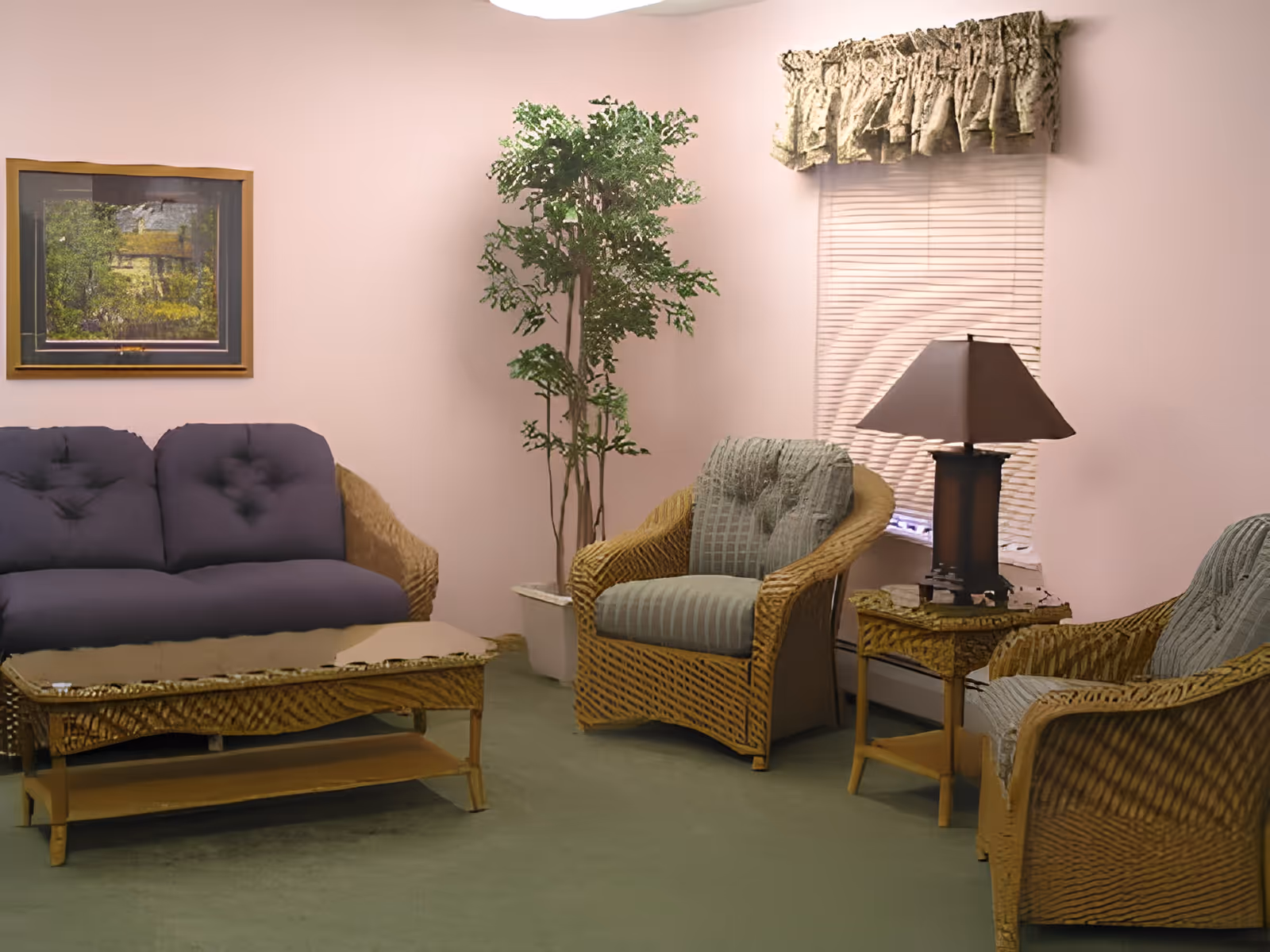 Cozy sitting area with wicker furniture including a purple sofa and two armchairs, a coffee table, potted plant, and a lamp by a window.