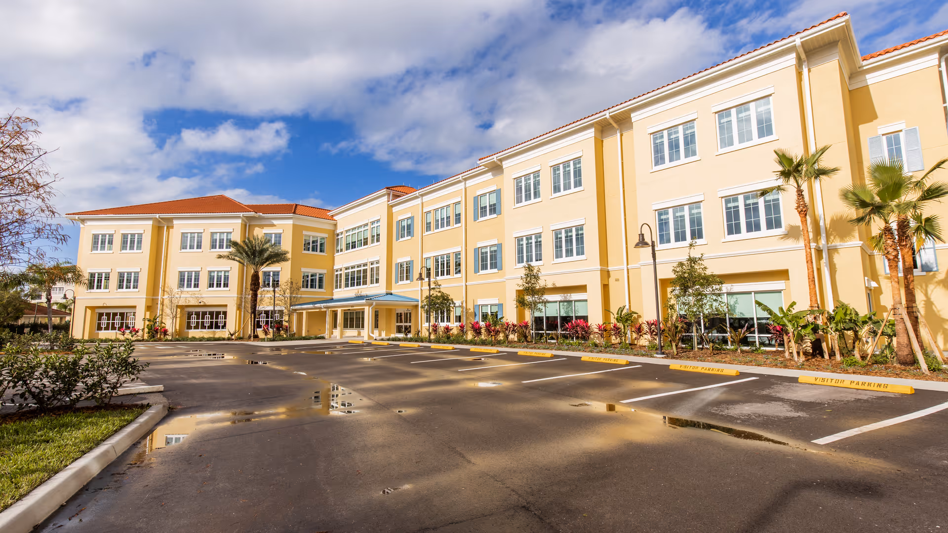 Exterior view of a three-story yellow building with multiple windows, palm trees, and a parking lot with marked visitor parking spaces under a partly cloudy blue sky.