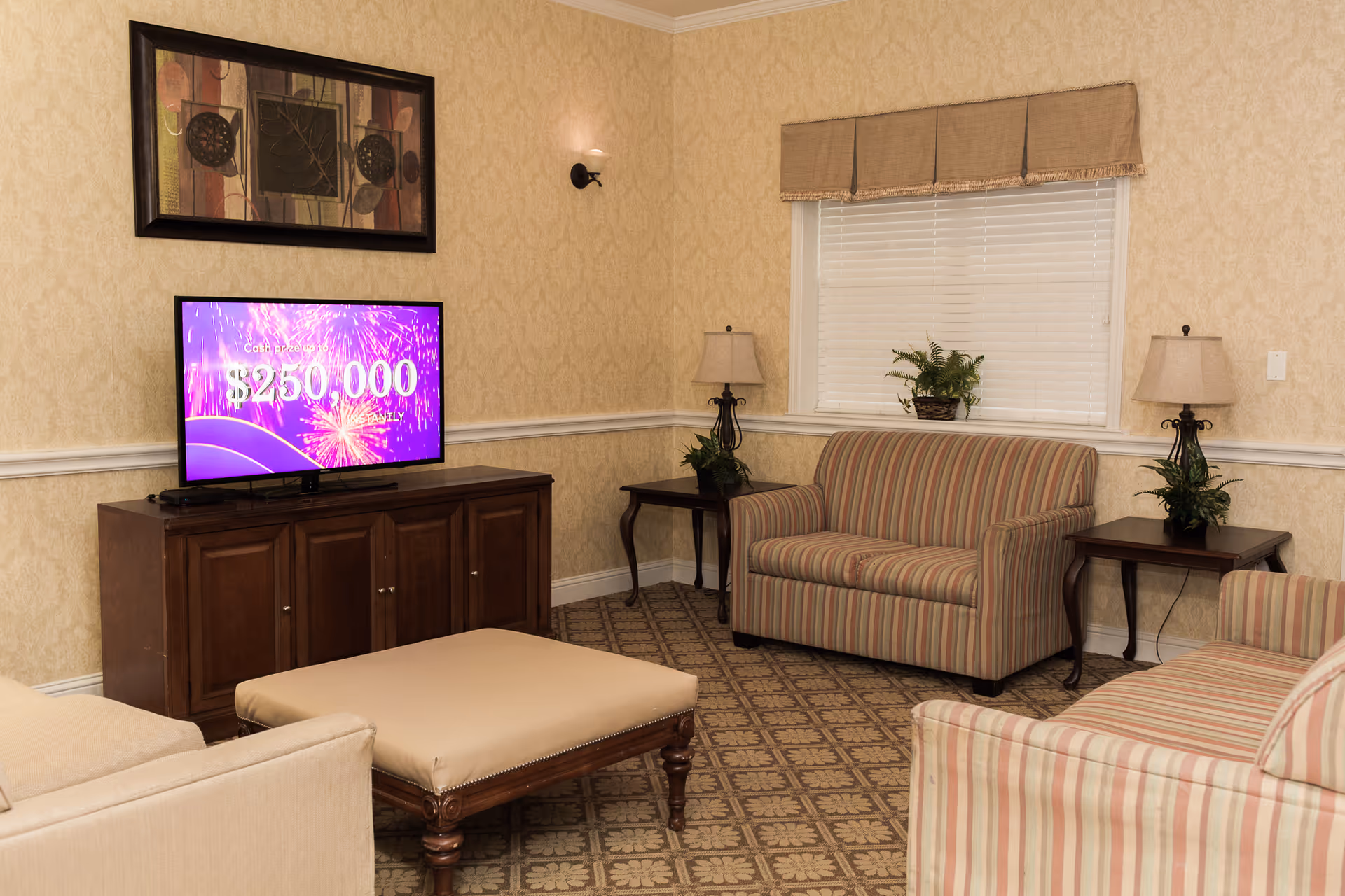 A cozy living room with patterned carpet, beige wallpaper, and a window with blinds and a valance. The room features a striped loveseat and matching armchair, a beige ottoman, two wooden side tables with lamps and plants, a wooden TV stand with a flat-screen TV displaying a purple screen with text about a $250,000 cash prize, and a framed abstract artwork on the wall.