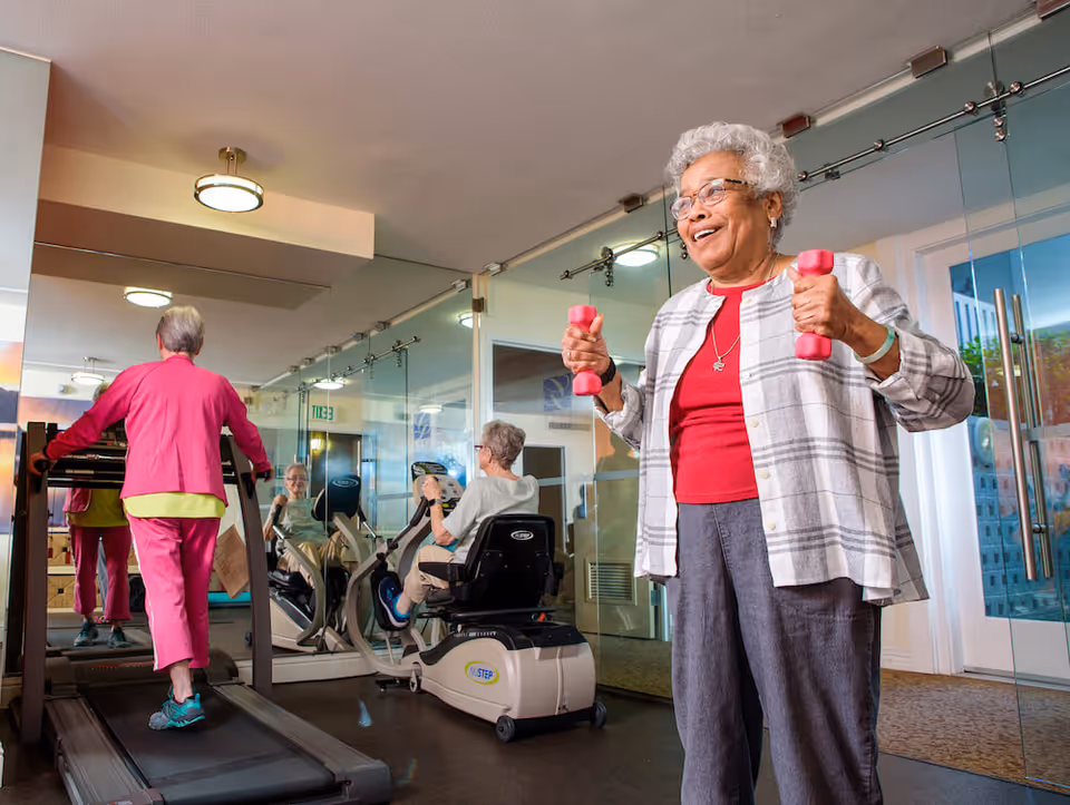 Two elderly women exercising in a fitness room. One woman is walking on a treadmill wearing a pink outfit, and the other woman is standing and lifting pink dumbbells, smiling. The room has large mirrors on the walls and glass doors.