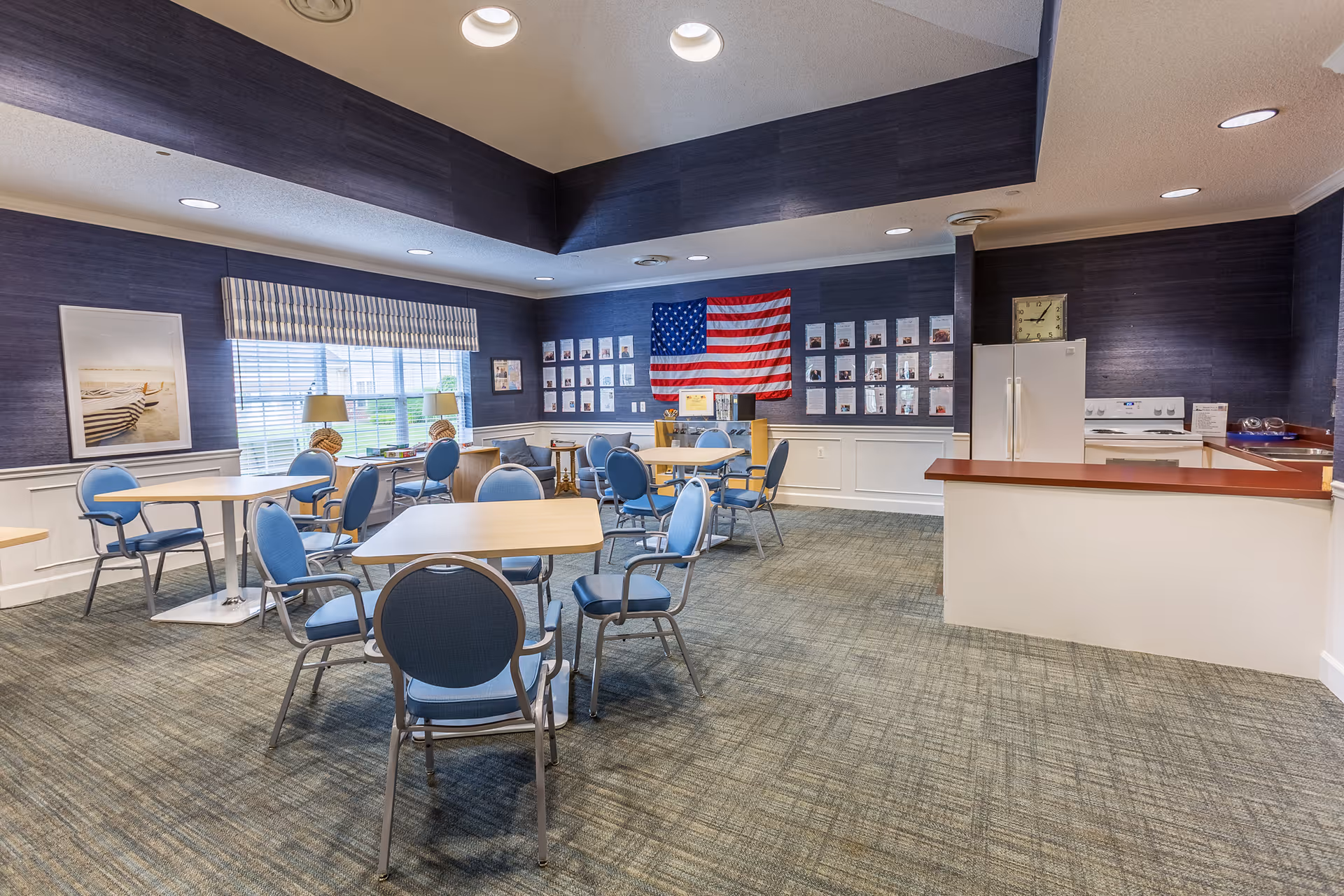 A communal dining area with several tables and blue cushioned chairs. The room has blue walls with white wainscoting, a large window with blinds, and an American flag displayed on the far wall above a bulletin board with various papers and photos. There is a small kitchen area with a white refrigerator, stove, and sink on the right side of the room. The carpet is patterned in shades of blue and green, and the ceiling has recessed lighting.