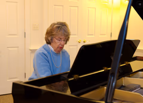 An elderly woman wearing glasses and a blue sweater is playing a black grand piano in a well-lit room with white double doors in the background.
