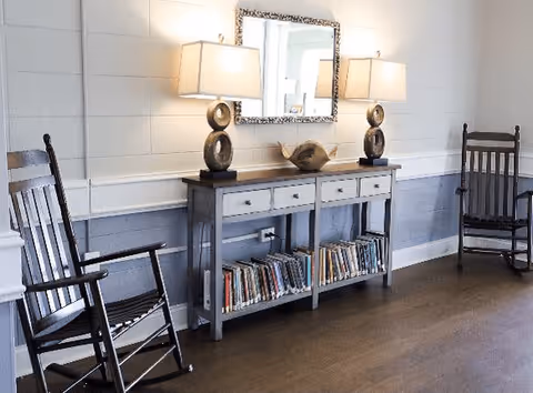 A cozy interior space featuring two dark wooden rocking chairs on either side of a gray console table with three drawers and a lower shelf filled with books. On top of the table are two decorative lamps with circular bases and a sculptural centerpiece. A rectangular mirror with a patterned frame hangs on the wall above the table. The room has light-colored walls with molding and a wooden floor.