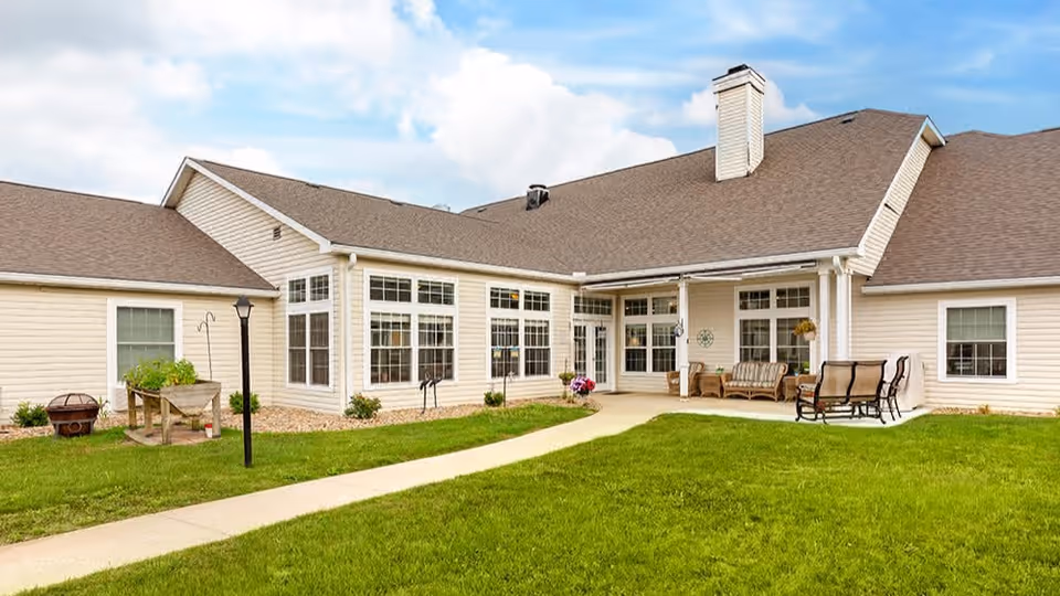 Exterior courtyard and patio of a single-story senior living building with large windows, a lawn, walkway, and outdoor seating.