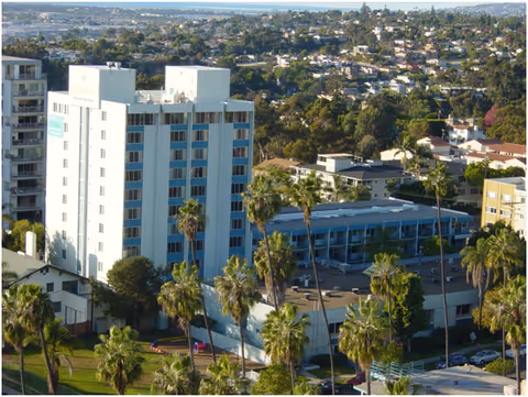 A multi-story white residential building with a low-rise annex surrounded by palm trees and a cityscape in the background.