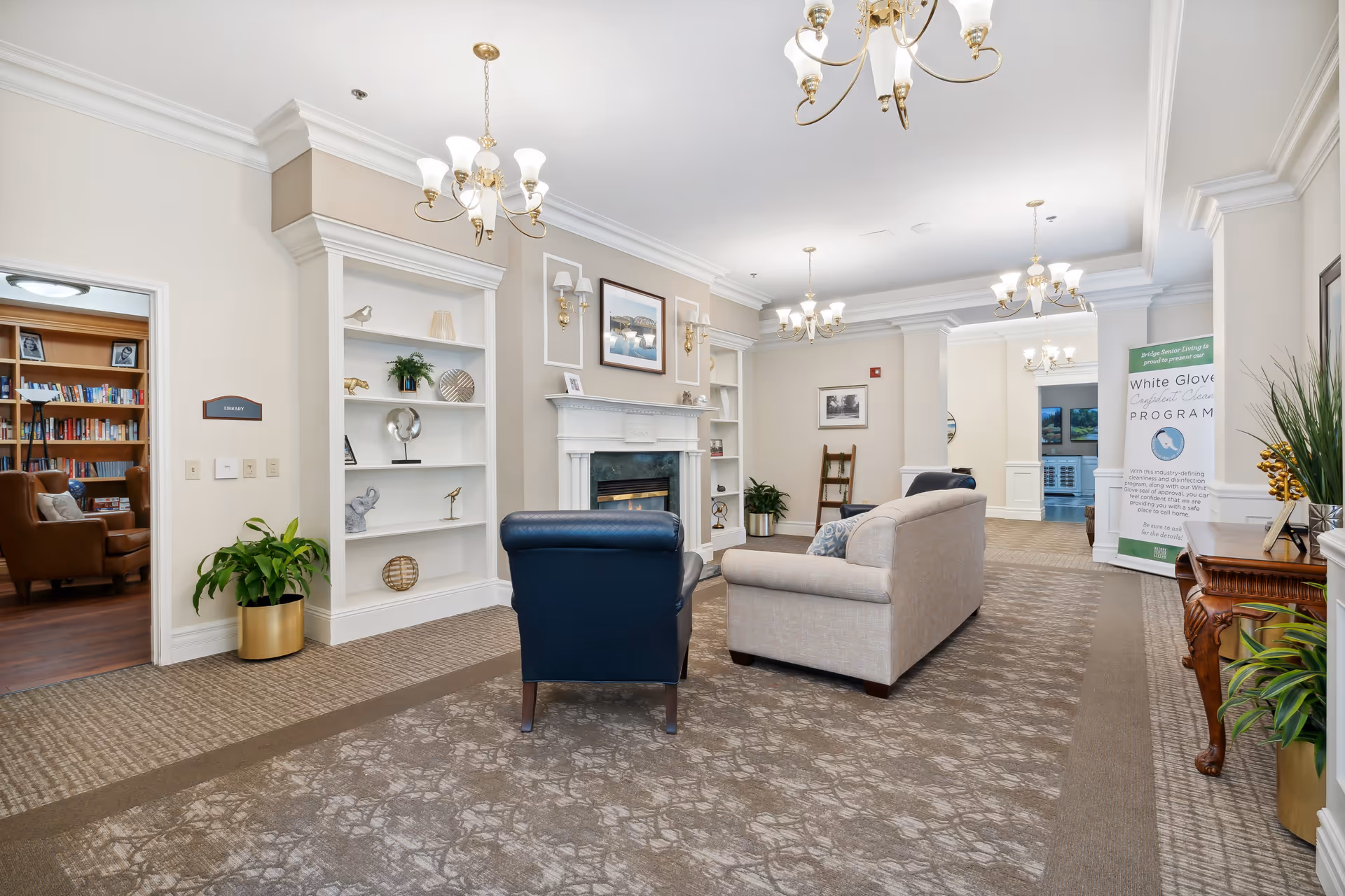 A well-lit assisted living facility common area with beige walls, carpeted floor, and white crown molding. The room features a beige sofa and a dark blue armchair facing a white fireplace with a framed picture above it. Built-in white shelves with decorative items flank the fireplace. To the left, an open doorway leads to a library with bookshelves and a brown leather chair. On the right side, there is a wooden console table with plants and a banner promoting a White Glove program. Multiple chandeliers hang from the ceiling.