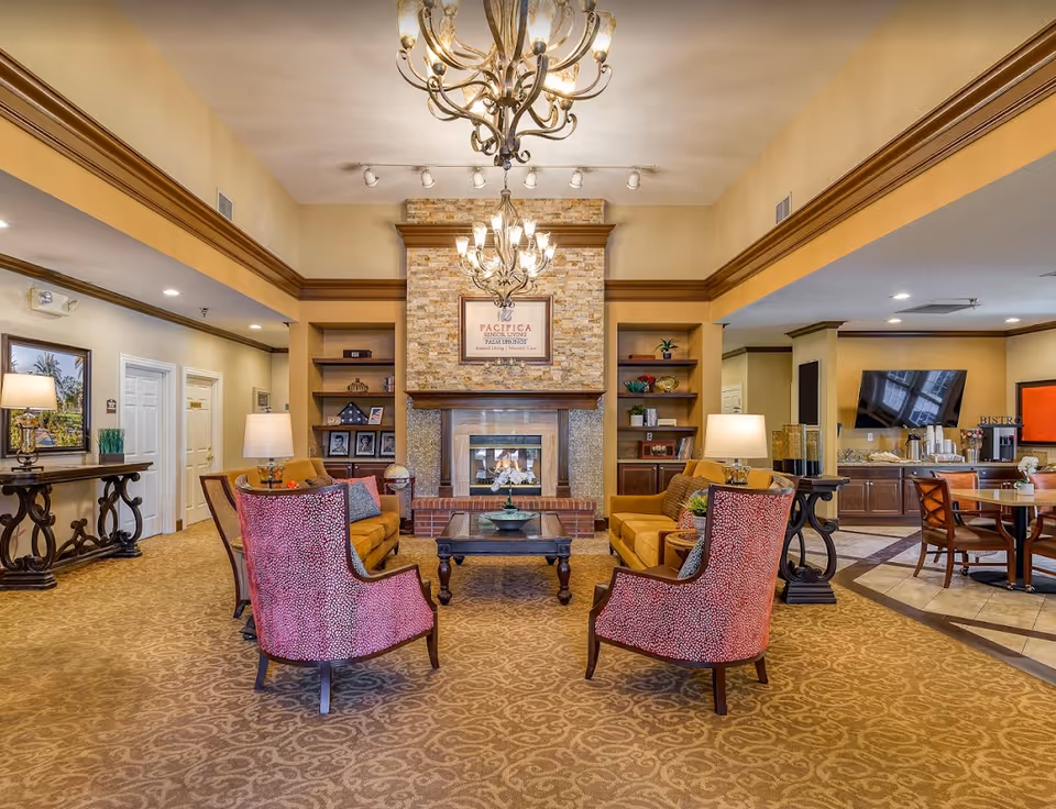 A cozy senior living facility common area with a fireplace in the center, flanked by built-in shelves. The room features two orange sofas and two red patterned armchairs arranged around a wooden coffee table. There are two chandeliers hanging from the ceiling, table lamps on side tables, and a television mounted on the wall in the background near a dining area with wooden chairs and tables.