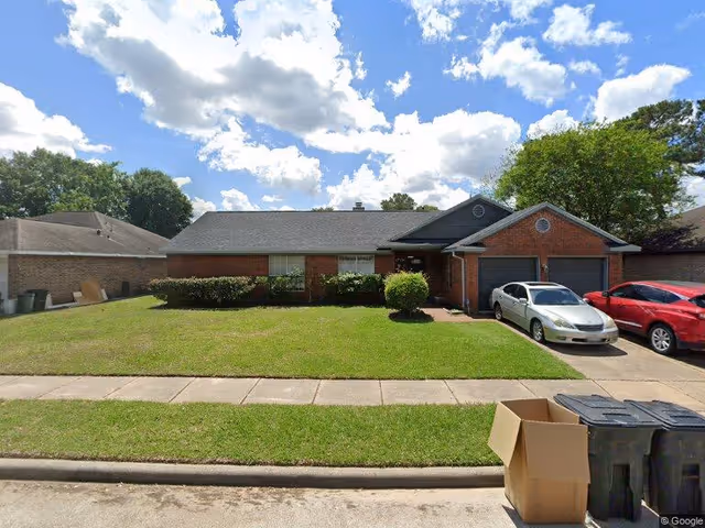 Front exterior view of a single-story brick house with a well-maintained lawn, two cars parked in the driveway, and a few trash bins and a cardboard box near the curb under a partly cloudy sky.
