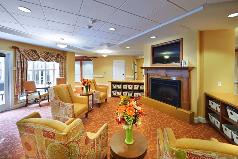 Bright communal lounge with patterned armchairs around small tables, a fireplace with a TV above, and storage cubbies.