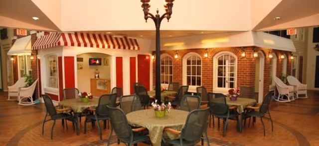 Indoor seating area with round tables covered with green tablecloths and black chairs arranged around them. The space features a decorative streetlamp in the center and facades resembling small storefronts with striped awnings and windows, creating a cozy, village-like atmosphere.