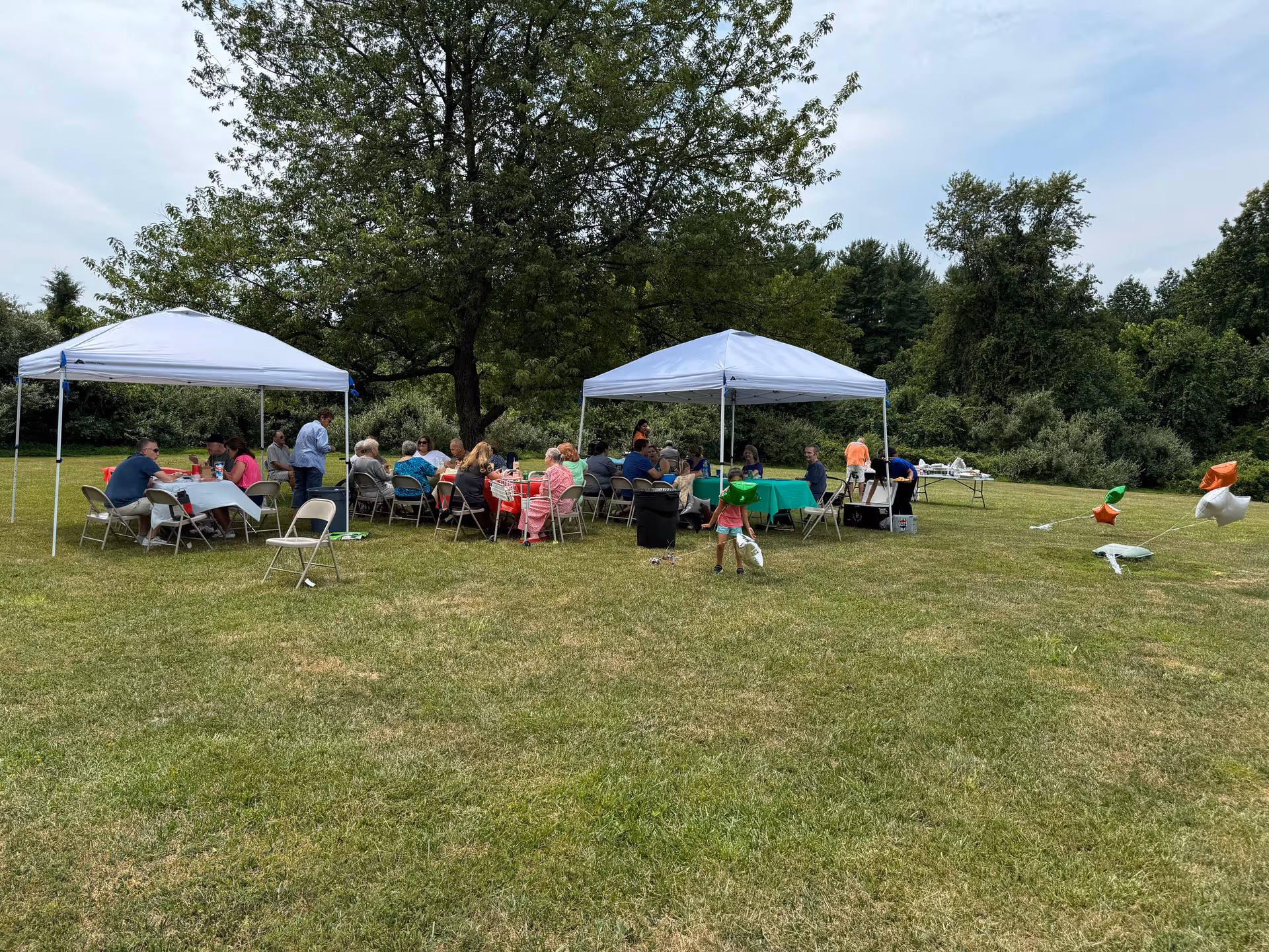 A group of people gathered outdoors under two white canopy tents on a grassy field, sitting at tables covered with red and green tablecloths. There are trees and bushes in the background, and some star-shaped balloons are tied to weights on the grass.
