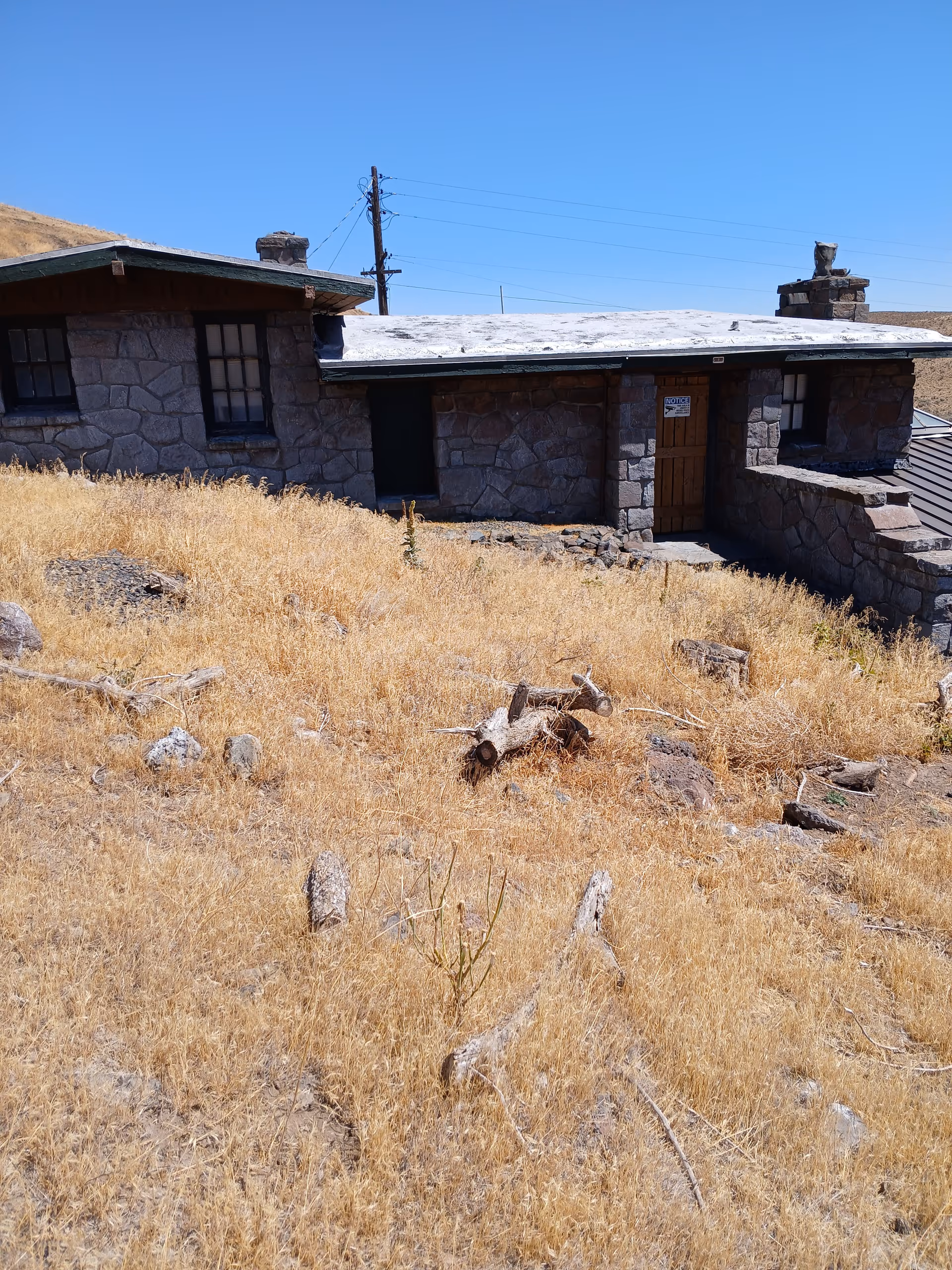 Low stone building with a wooden door behind a dry grassy hillside under a clear blue sky.