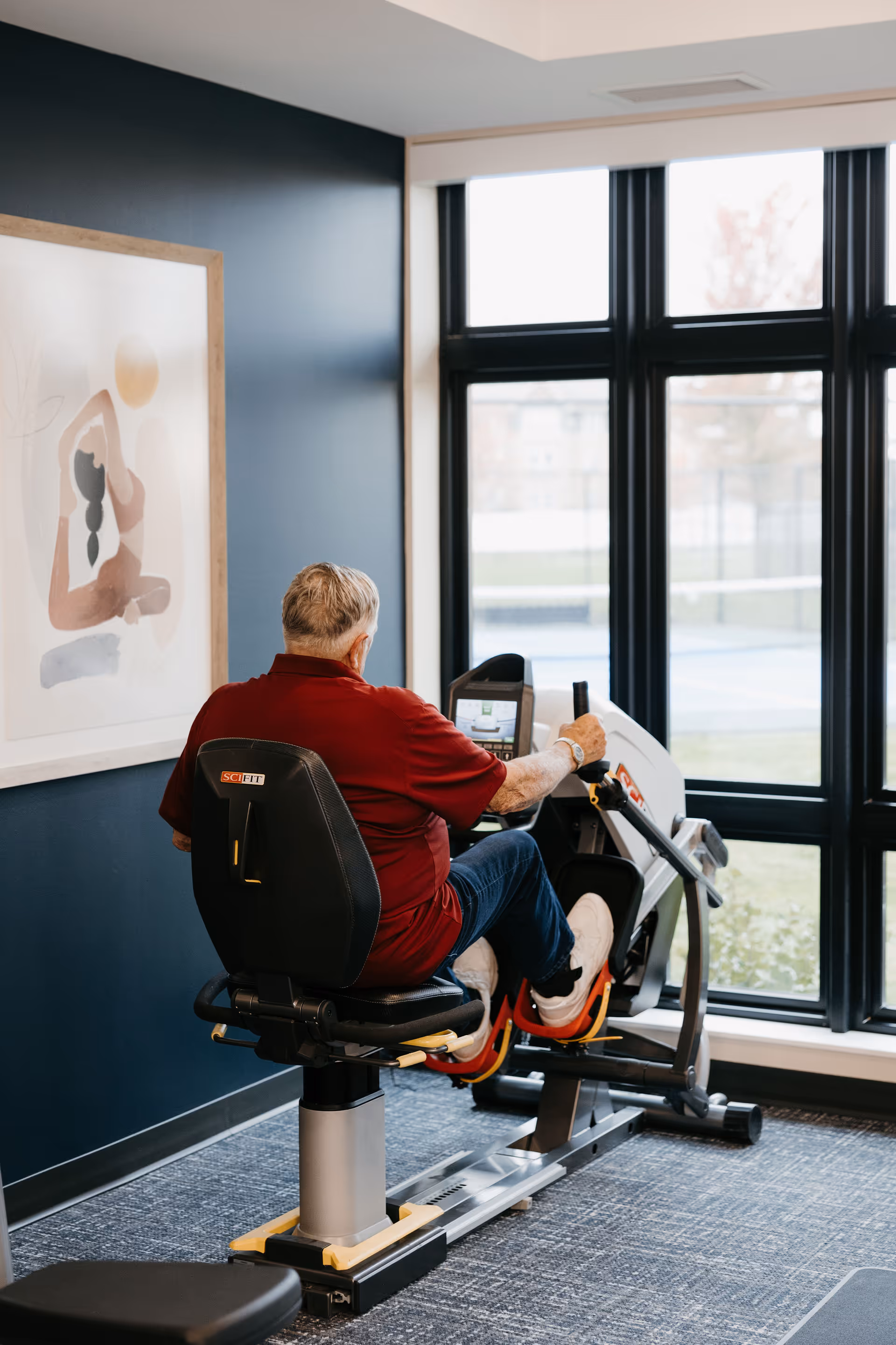 An elderly man wearing a red shirt and jeans is exercising on a seated leg press machine in a fitness room with large windows and a framed abstract artwork on the wall.