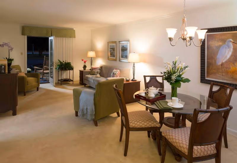 A cozy living room and dining area in a retirement community. The living room features a sofa with floral cushions, two armchairs, side tables with lamps, and framed artwork on the wall. The dining area has a round glass table with four wooden chairs, a vase with white flowers, and a tea set. A chandelier hangs above the dining table. The room is warmly lit and has an open door leading outside to a patio with rocking chairs.
