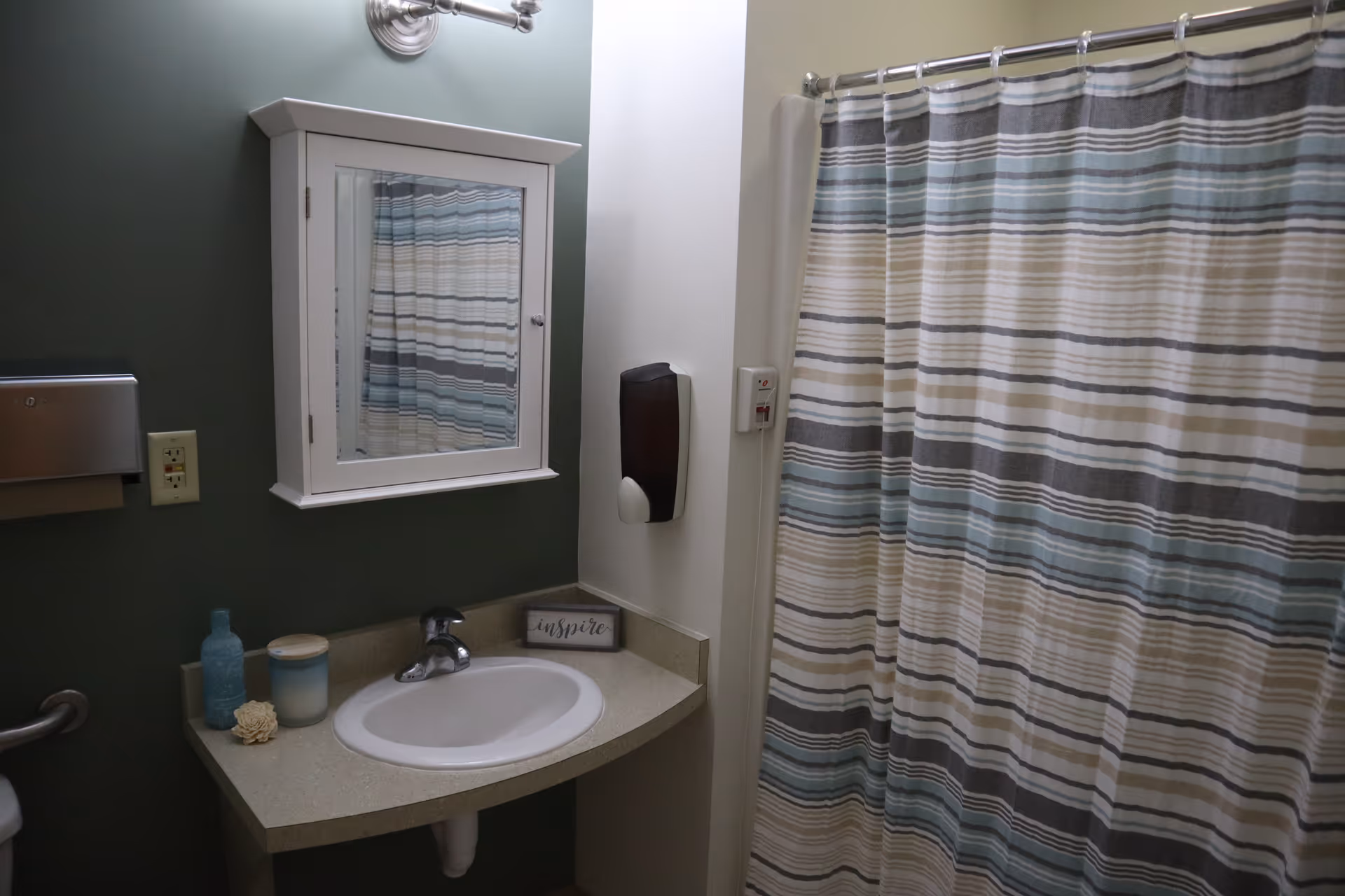 Bathroom with a white sink and countertop, a wall-mounted white medicine cabinet with a mirror, a striped shower curtain in shades of blue, gray, and beige, a soap dispenser on the wall, and decorative items including a blue bottle, a candle, and a small sign that says 'inspire'.