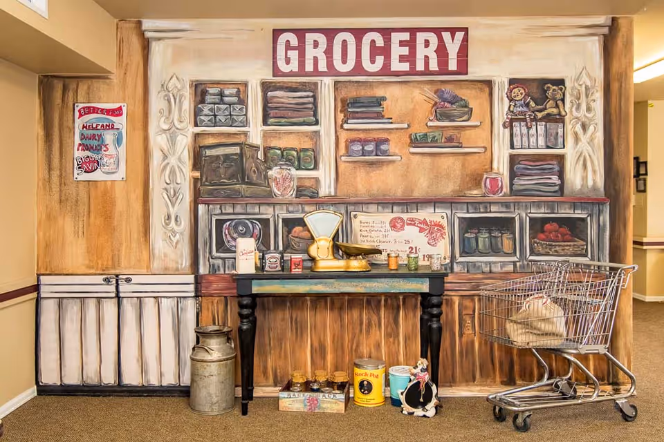 A mural painted on a wall depicting a vintage grocery store scene with shelves holding various items like canned goods, folded cloths, and toys. In front of the mural is a small black table with a vintage scale and other small items. To the right is a shopping cart with a bag inside. The setting appears to be indoors with beige walls and carpeted floor.