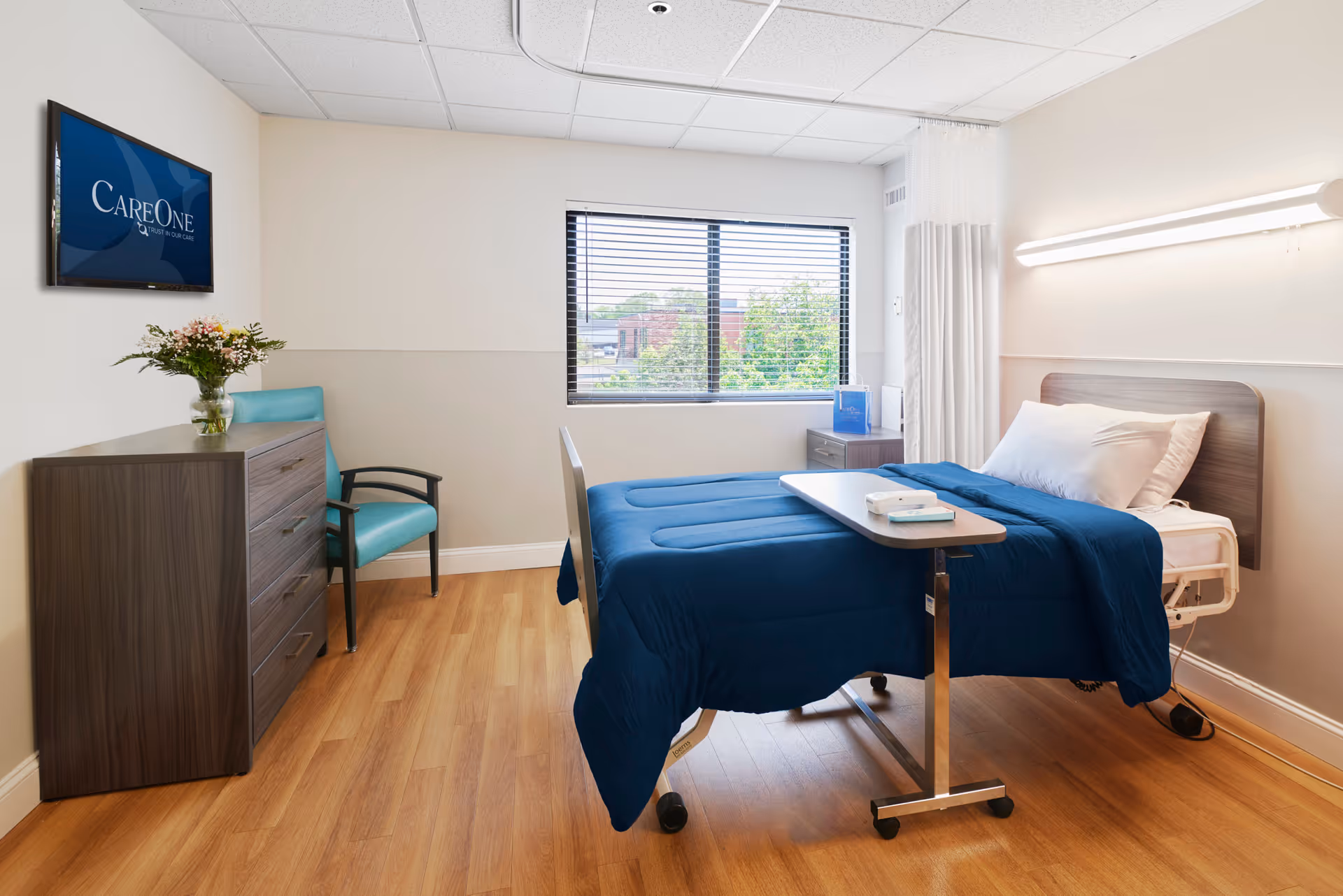 A clean and bright patient room with a hospital bed covered in a blue blanket, a small overbed table, a wooden dresser with a vase of flowers, a teal armchair, a window with blinds showing greenery outside, and a wall-mounted TV displaying the CareOne logo.