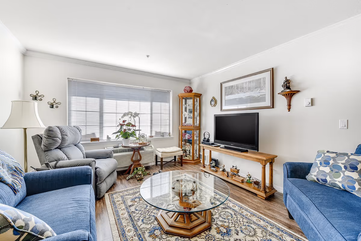 A bright living room with a large window covered by blinds, a gray recliner chair, a blue sofa with patterned pillows, a glass coffee table on a decorative rug, a wooden TV stand with a flat-screen TV, and a corner display cabinet with glass doors containing various items. The walls are white with a framed picture and small decorative shelves.