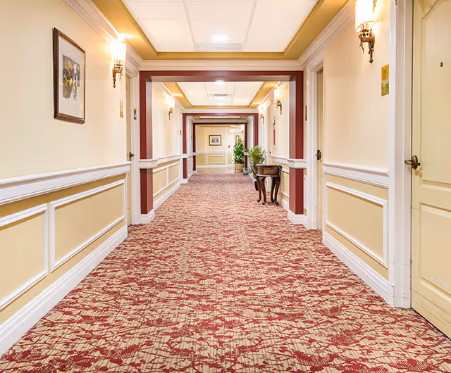 A well-lit, carpeted hallway in a senior living facility with cream-colored walls, white trim, and maroon accents. The hallway features wall sconces, framed artwork, a small table with plants, and multiple doors along the sides.