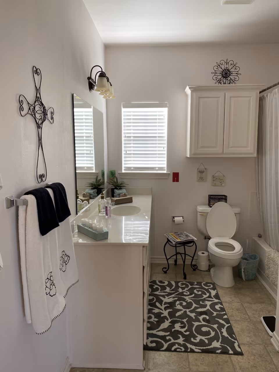 Bright bathroom with a single-sink vanity and mirror, window with blinds, wall cabinet, toilet, shower and decorative accents.