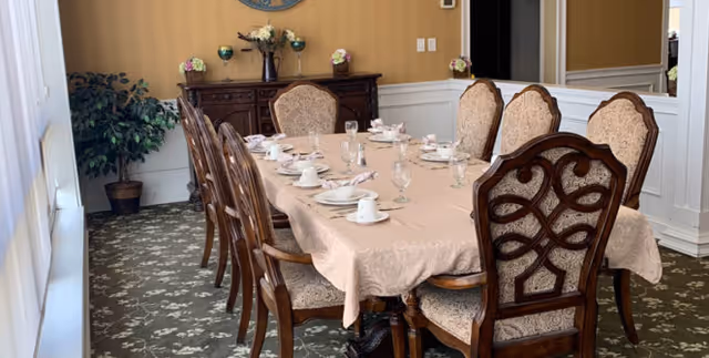A formal dining room with a long rectangular table covered with a beige tablecloth, set with white cups, saucers, plates, and glasses. The table is surrounded by eight ornate wooden chairs with upholstered seats and backs. In the background, there is a wooden sideboard with decorative items and a potted plant in the corner. The room has a patterned carpet and light-colored walls with wainscoting.