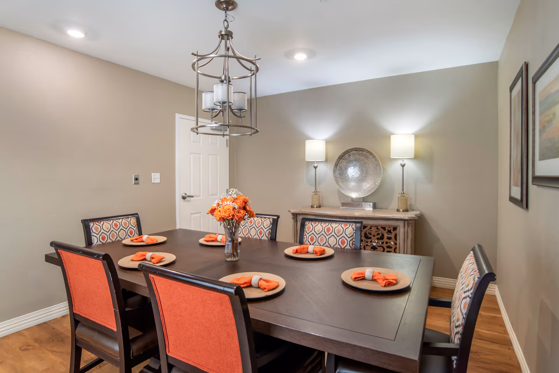 A dining room with a dark wooden table set for six, each place having a round placemat and an orange napkin with a white napkin ring. The chairs have patterned backs and orange fabric on the backrests. A vase with orange flowers is centered on the table. The room has beige walls, two table lamps on a sideboard with a decorative silver bowl between them, and two framed pictures on the right wall. A modern chandelier hangs above the table.