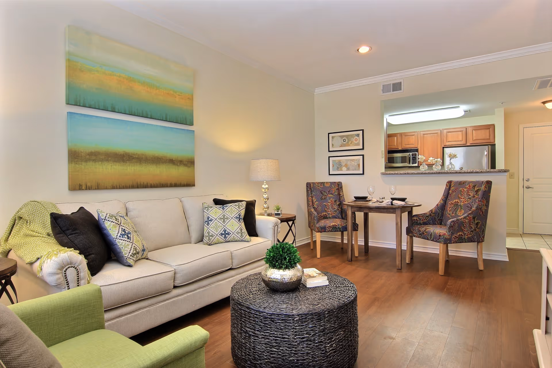A cozy living room area with a beige sofa adorned with patterned pillows and a green throw blanket. In front of the sofa is a round woven coffee table with a small potted plant and a book. To the right, there is a small dining area with a round wooden table set for two and two upholstered chairs with a floral pattern. The background shows a kitchen with wooden cabinets, a microwave, and a refrigerator. The walls are decorated with two abstract paintings above the sofa and two framed pictures near the dining area. The floor is wooden, and the room is softly lit with a table lamp and recessed ceiling lights.