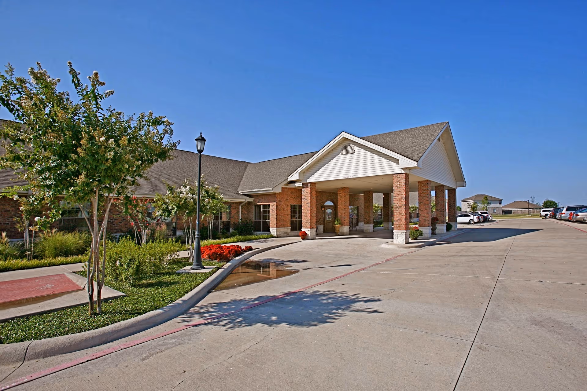 Exterior view of Royse City Medical Lodge showing the building entrance with a covered drop-off area supported by brick columns, a driveway, landscaped greenery with trees and flowers, and parked cars under a clear blue sky.