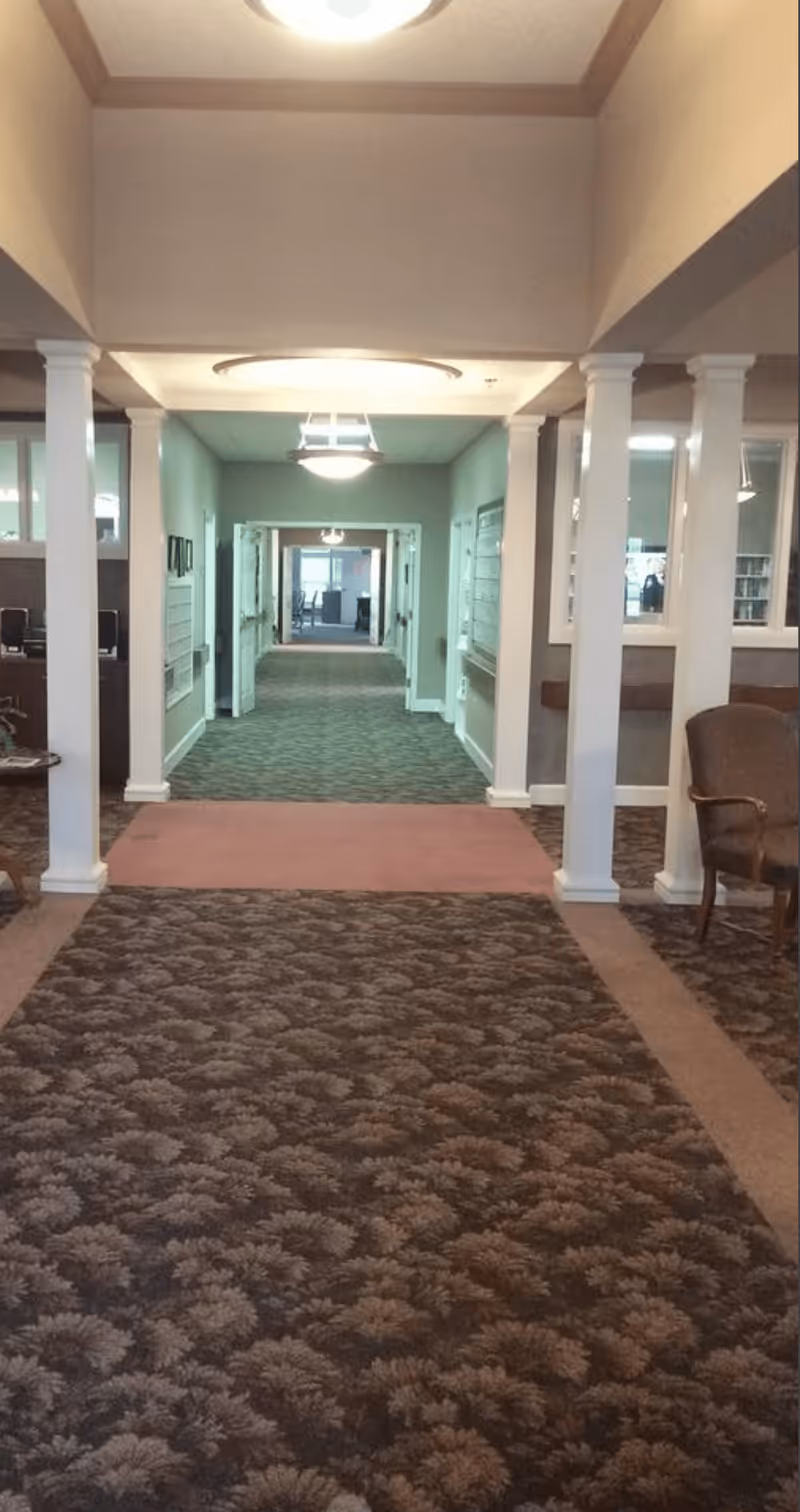 Interior hallway of a senior living facility with patterned carpet, white columns, beige walls, and ceiling lights. There are chairs and windows on the right side, and the hallway extends into another room in the distance.