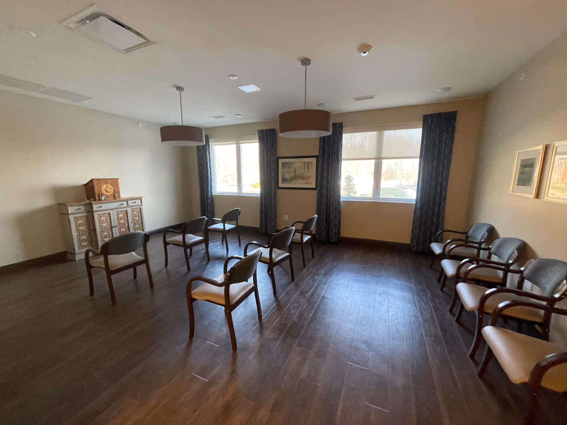 A spacious room with wooden flooring and several chairs arranged in a semi-circle and along the walls. The room has two large windows with dark curtains, two ceiling pendant lights, and framed artwork on the walls. There is a decorative cabinet against one wall.