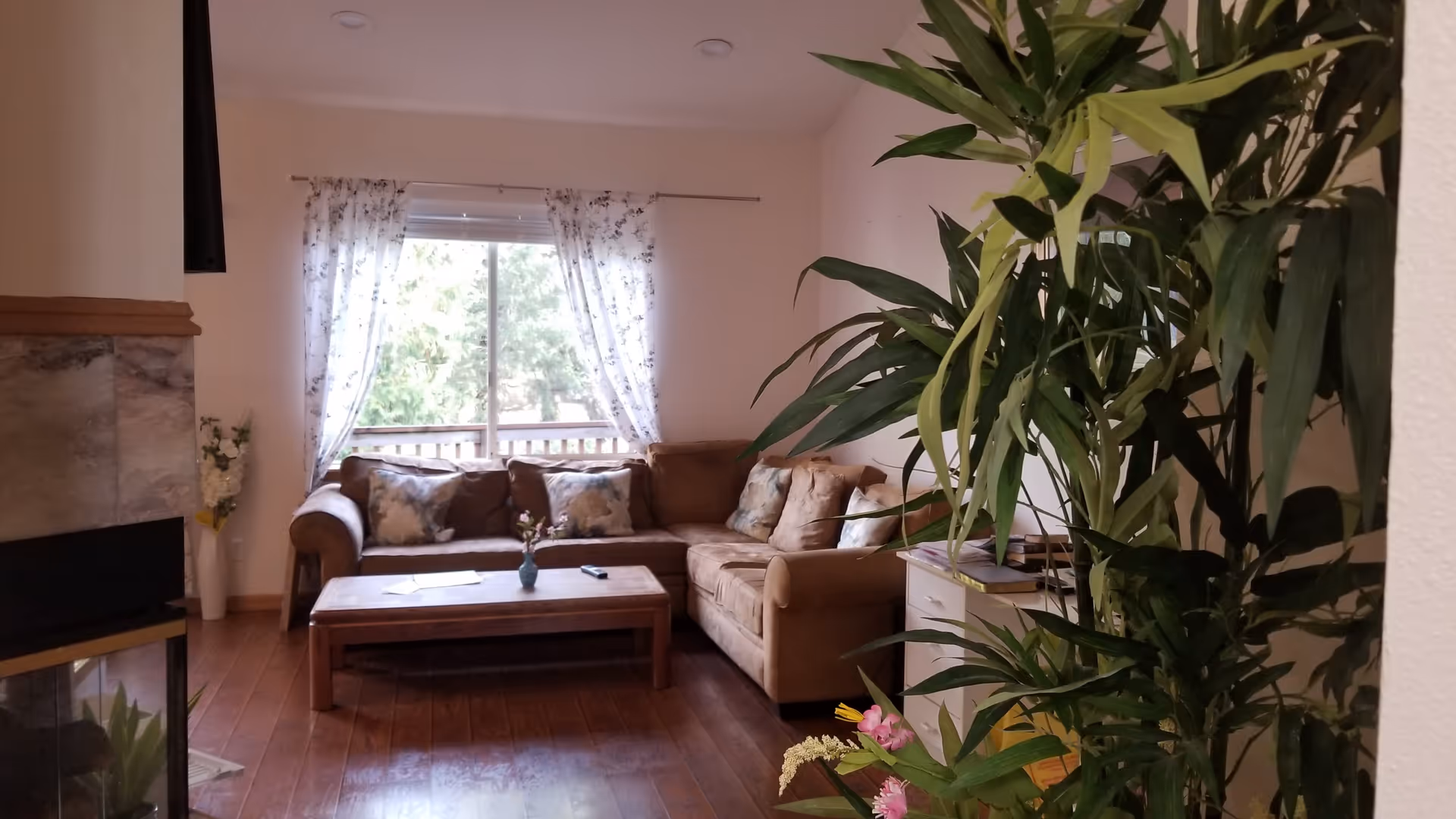 Cozy living room with an L-shaped tan sofa, wooden coffee table, large window with floral curtains, and indoor plants on a hardwood floor.