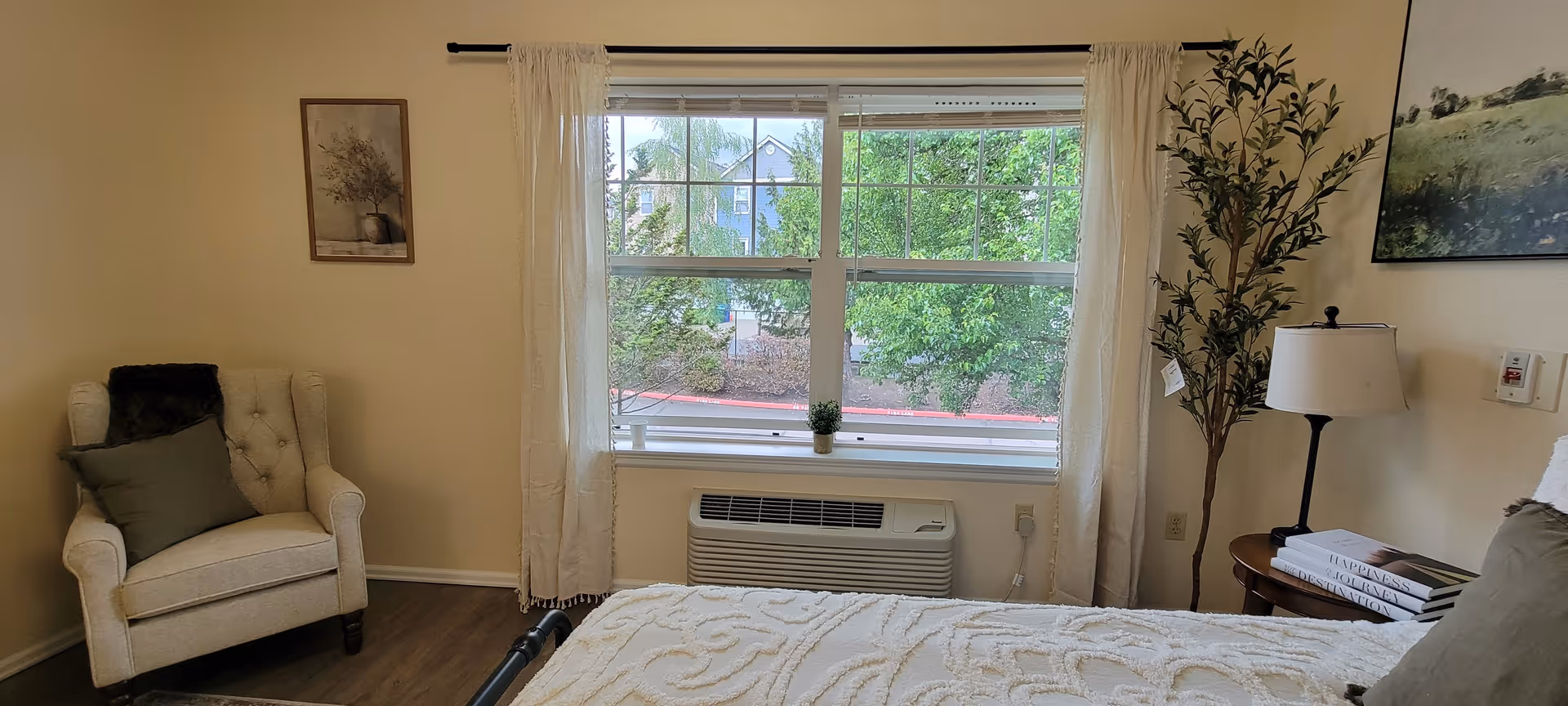 A cozy bedroom with a large window showing green trees outside. The room features a beige armchair with a dark pillow and throw, a bed with a textured white blanket, a small round wooden side table with a lamp and stacked books, and a tall potted plant. Light curtains frame the window, and a framed picture hangs on the wall.