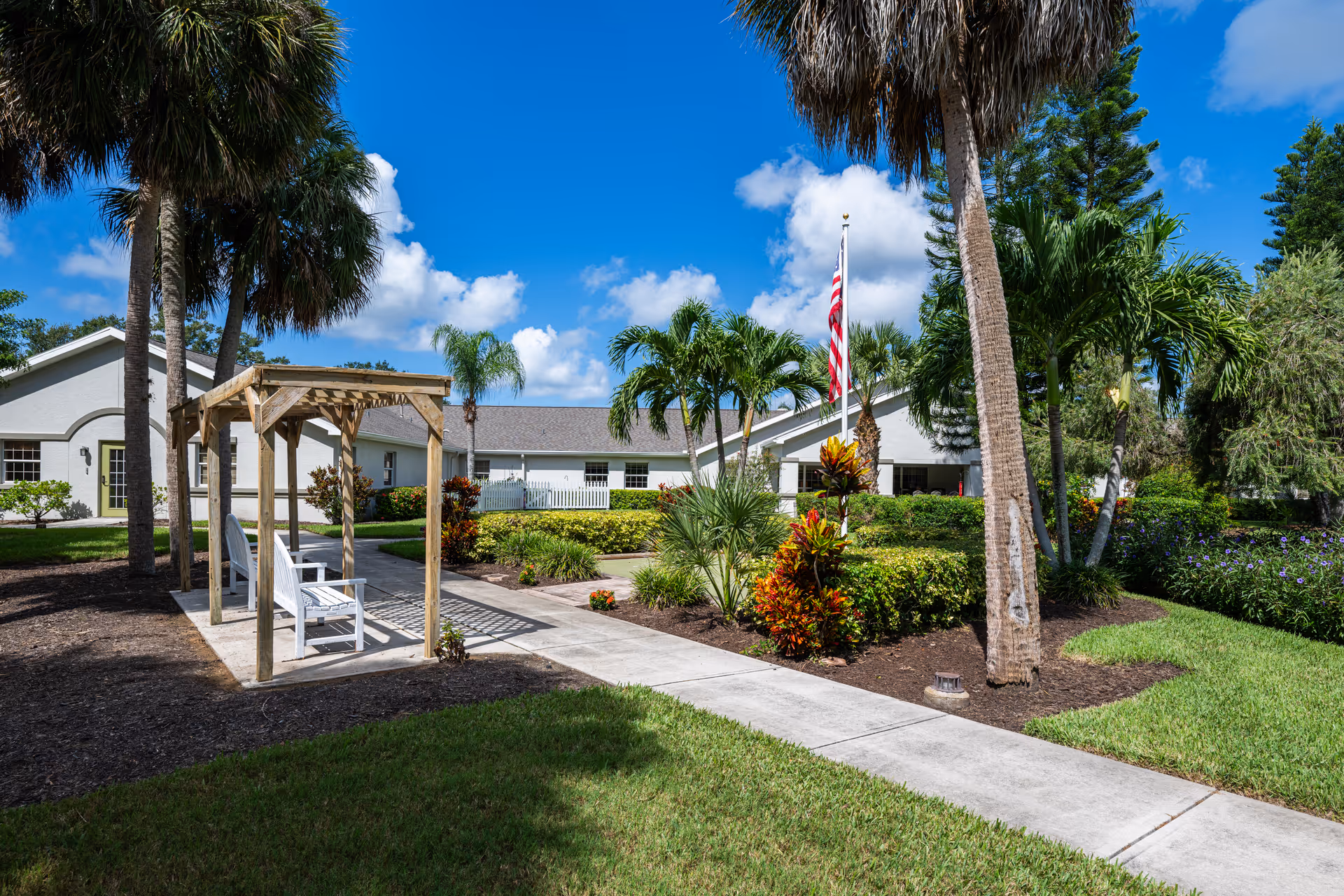 Sunny landscaped courtyard with palm trees, a wooden pergola with white benches, an American flag, and the facility building in the background.