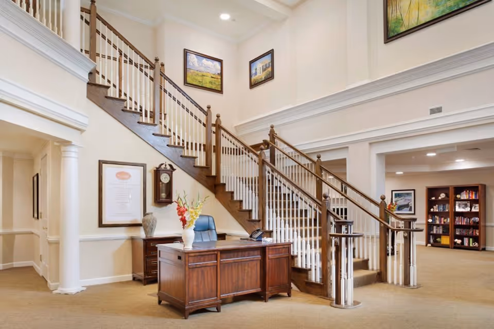 Interior view of a senior living facility lobby area featuring a wooden staircase with white balusters, a wooden desk with a vase of flowers and a chair, framed artwork on the walls, a wall clock, and a bookshelf in the background.
