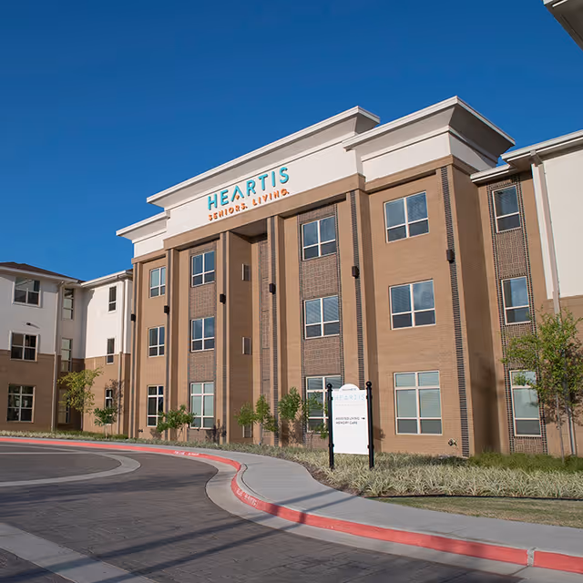 Exterior view of the Heartis Seniors Living building under a clear blue sky, featuring a multi-story brick and beige facade with multiple windows and a curved driveway in front.