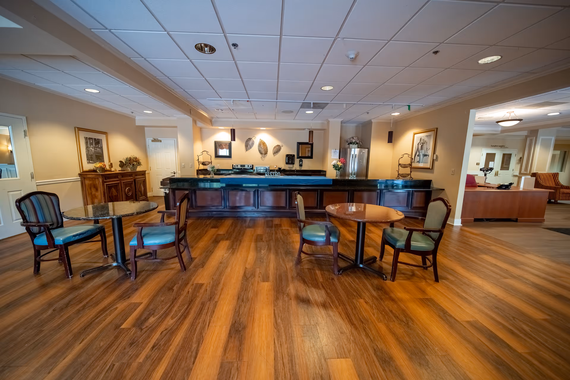 Spacious senior living common dining area with small round tables and chairs and a long service counter/kitchen in the background.