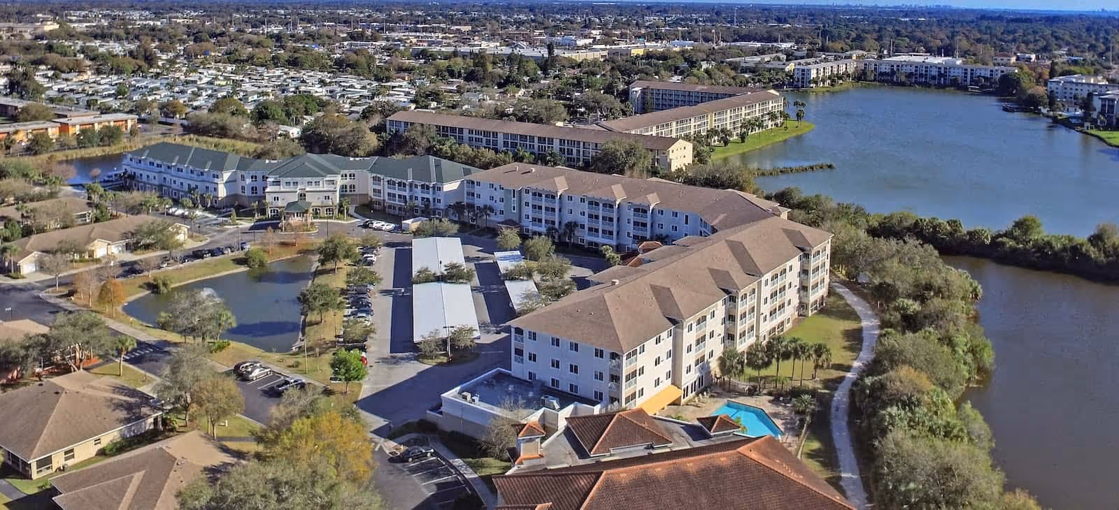 Aerial view of a senior living facility named Water's Edge of Bradenton, showing multiple residential buildings surrounding a large pond and a swimming pool. The area includes parking lots, trees, and walking paths, with a suburban landscape in the background.