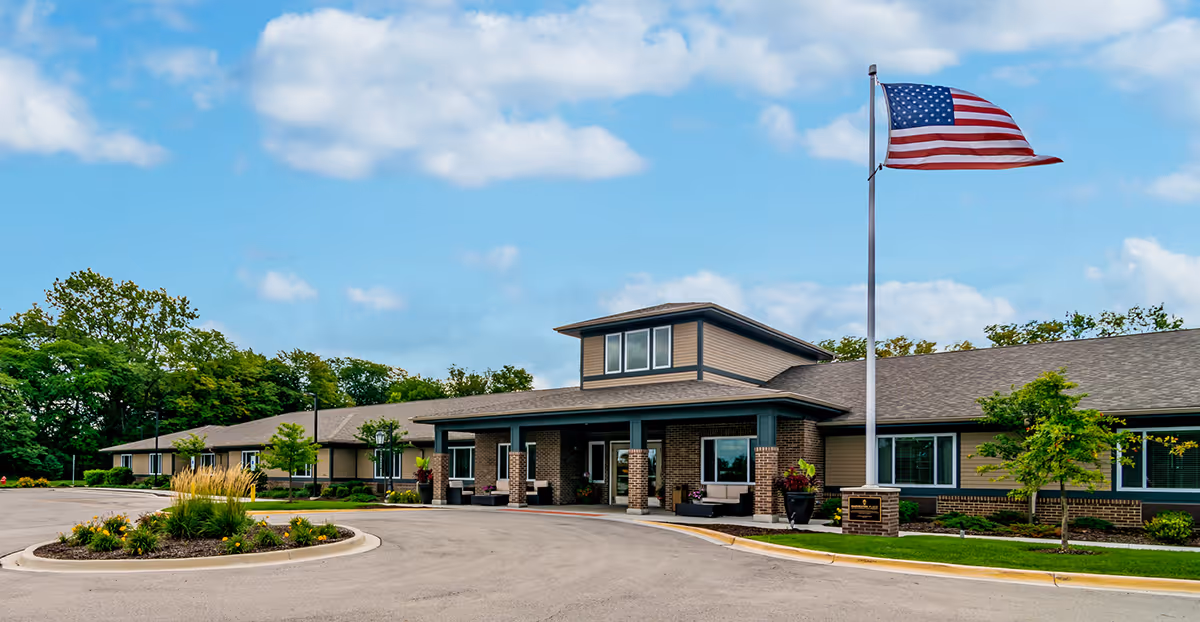 Front entrance of a single-story senior living building with an American flag, circular driveway, and landscaped beds.