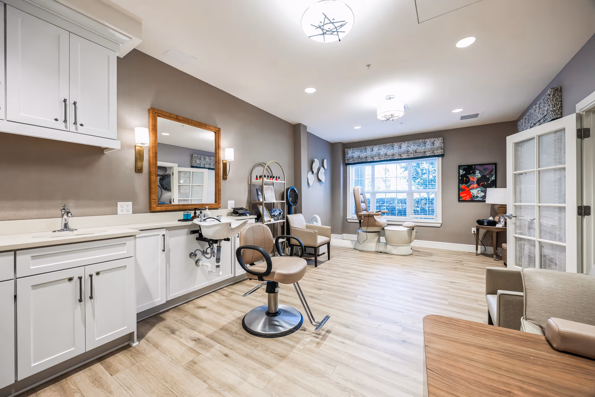 A bright salon-style room with styling chairs, a wash basin, cabinets, and seating by a large window.