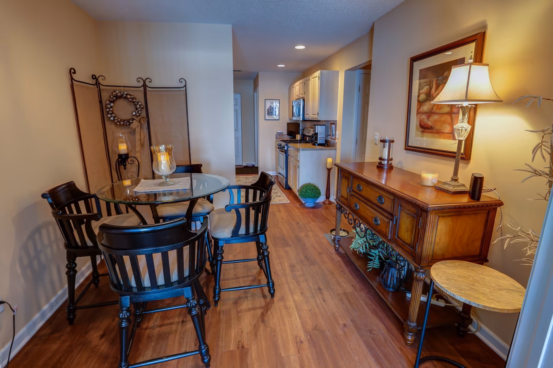 Interior view of a dining area with a round glass table surrounded by four black wooden chairs. To the right, there is a wooden sideboard with a lamp, candles, and a framed artwork above it. The room has wooden flooring and leads into a kitchen area with white cabinets and appliances.