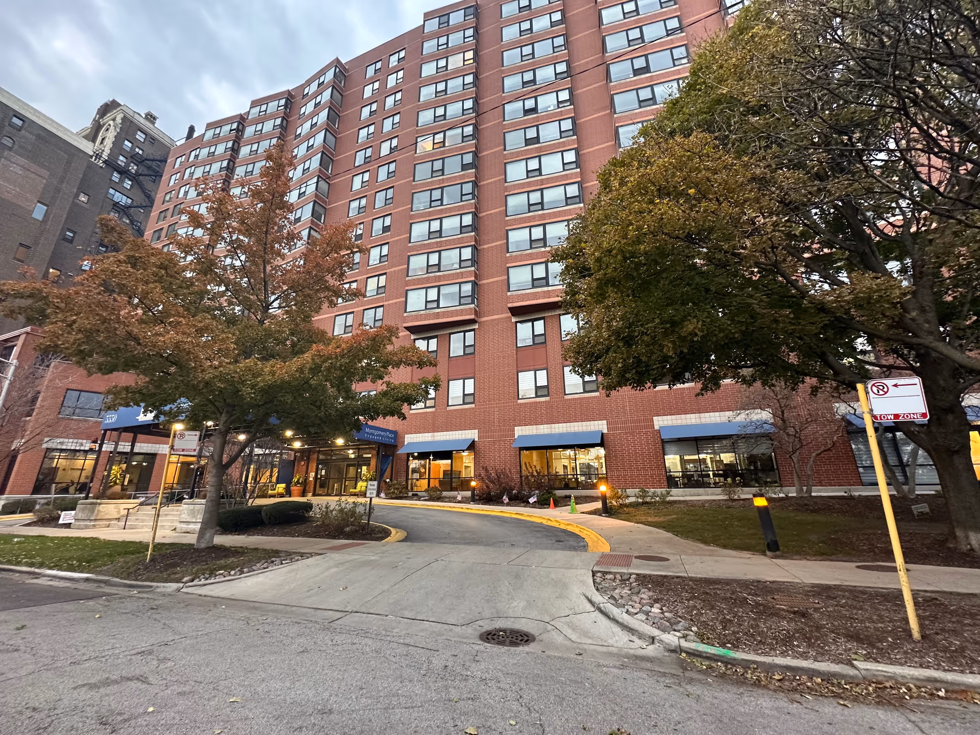 Exterior view of a tall brick building with multiple windows, trees with autumn foliage in front, and a curved driveway leading to the entrance of Montgomery Place Retirement Community.