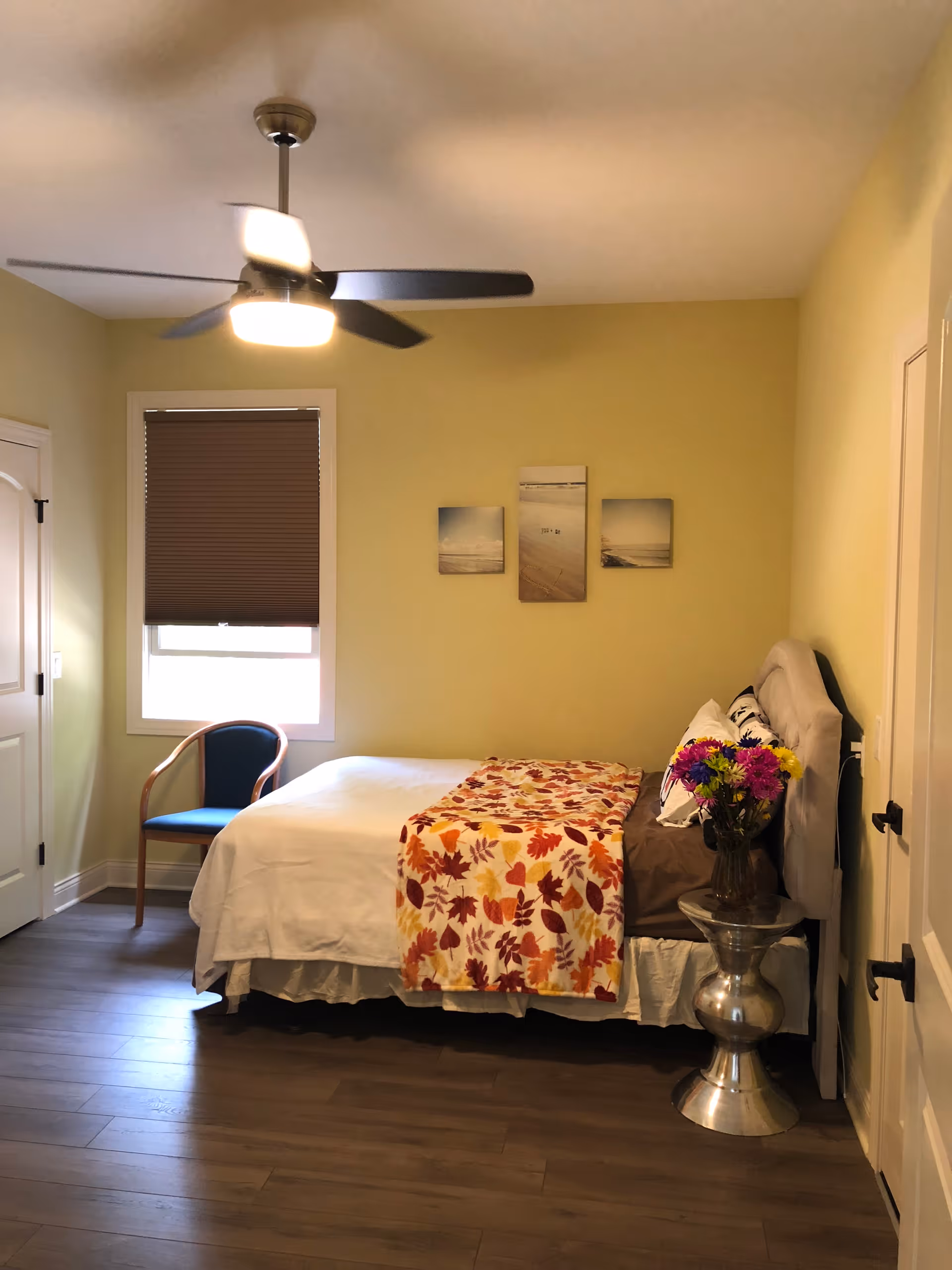 A cozy bedroom with a bed covered in white sheets and a colorful autumn leaf patterned blanket. There is a small side table with a vase of flowers next to the bed. A blue chair is placed near a window with a brown blind. The walls are painted light yellow, and there are three small framed pictures hanging above the bed. A ceiling fan with a light is mounted on the ceiling.