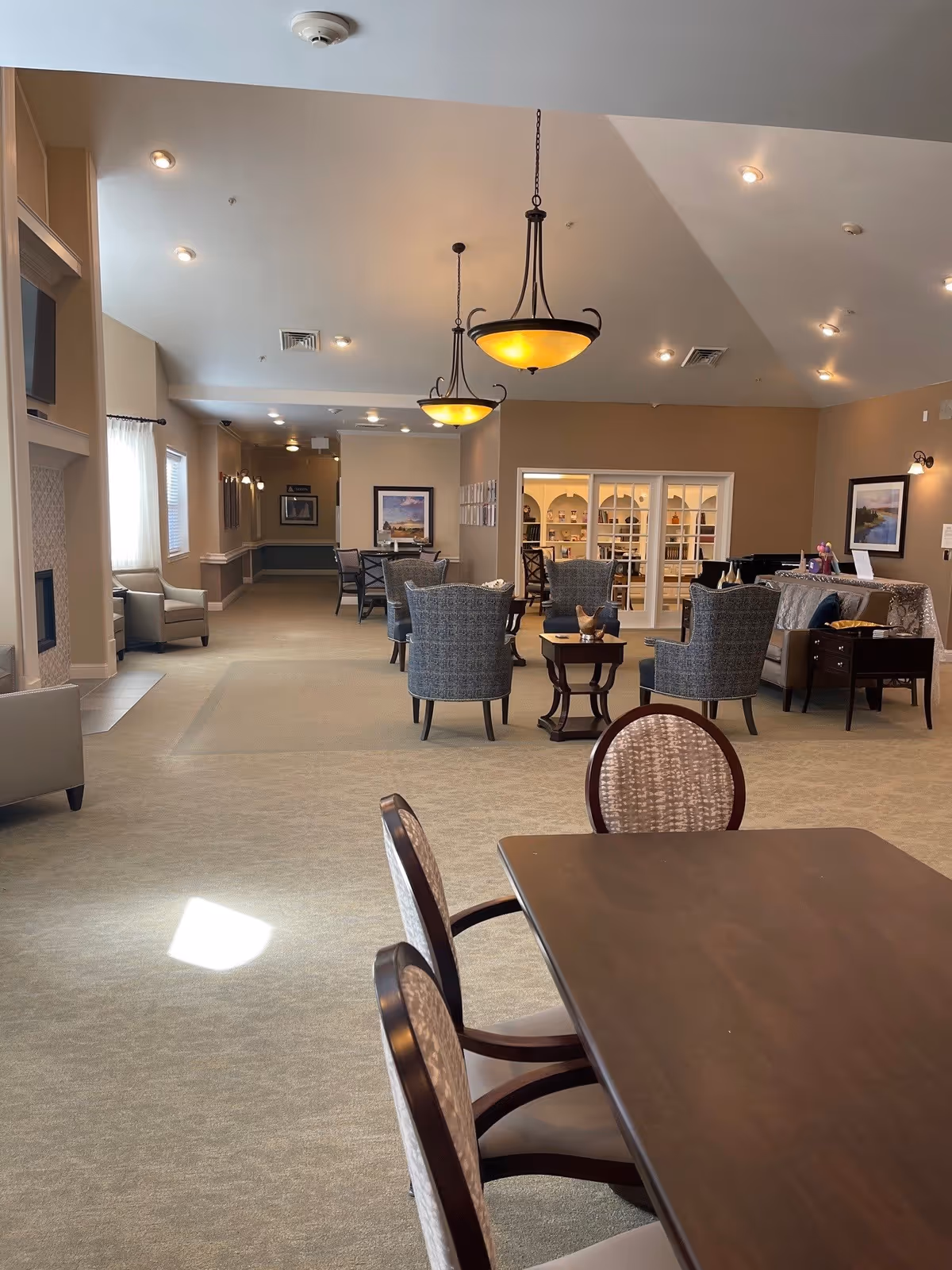 Spacious senior living common area with a dining table in the foreground, upholstered chairs and lounge seating under hanging chandeliers.