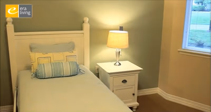 A small bedroom with a single bed featuring a white wooden headboard, light blue bedding, and decorative pillows. Next to the bed is a white nightstand with a drawer and a lamp with a beige lampshade. There is a window with blinds on the right side, showing a view of green grass outside.